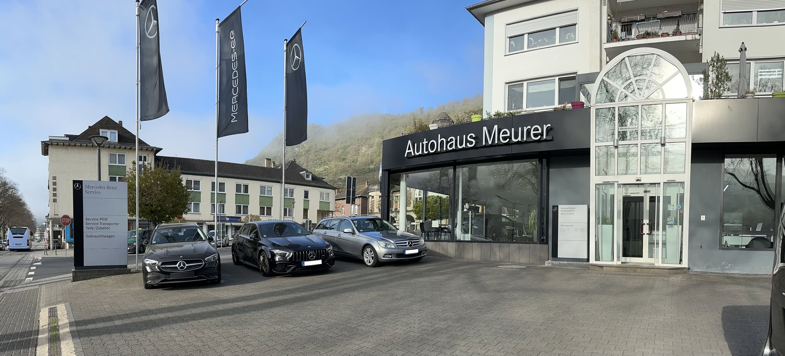 Außenansicht des Autohauses ‚Autohaus Meurer‘ mit drei geparkten Mercedes-Benz-Fahrzeugen davor. Das Gebäude hat große Glasfenster und ein modernes Design. Zwei hohe Fahnen mit Mercedes-Benz-Logo und -Schriftzug stehen vor dem Gebäude. Ein Schild am Eingang weist auf folgende Dienstleistungen hin: ‚Service-Direkt-Annahme‘, ‚Teile & Zubehör‘ und ‚Lackiererei‘. Im Hintergrund sind weitere Gebäude, Bäume und ein Hügel zu sehen.
